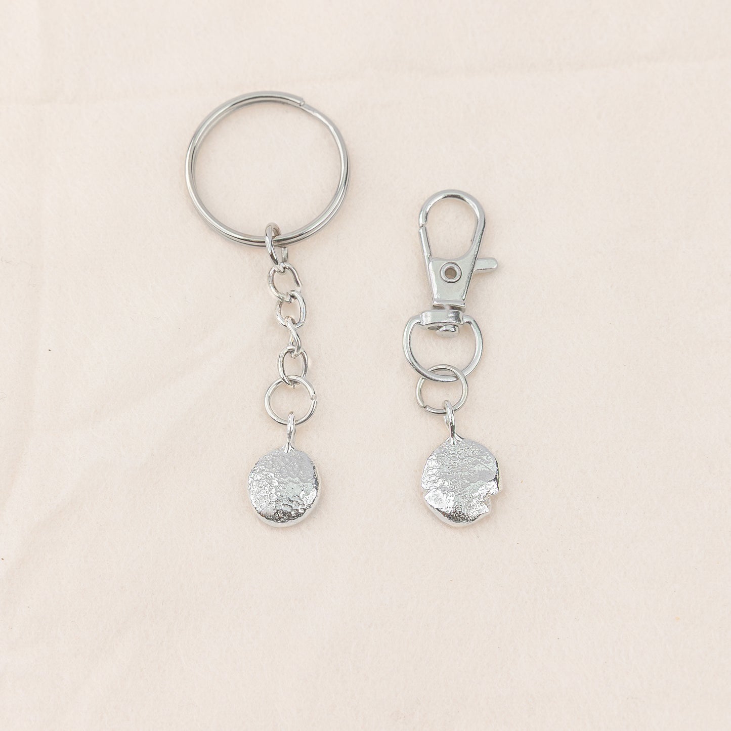 Two silver keychains with textured round pendants on a beige background