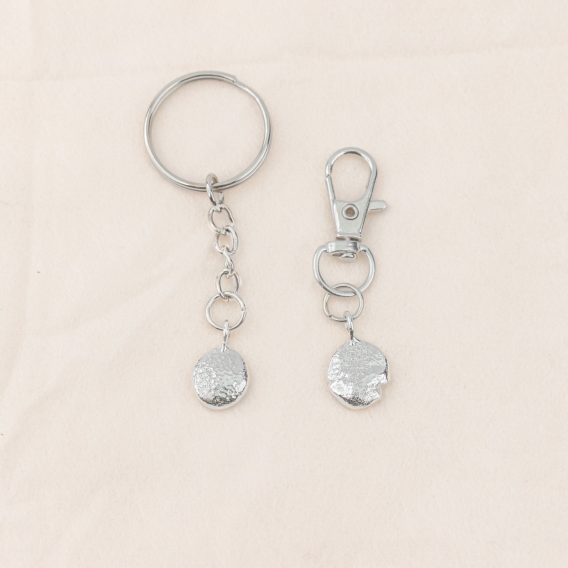 Two silver keychains with textured round pendants on a beige background