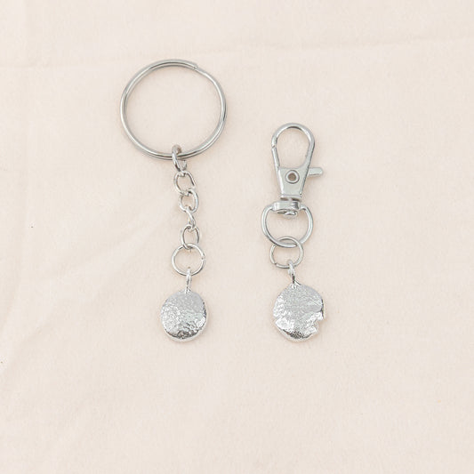 Two silver keychains with textured round pendants on a beige background